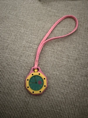 Färgglad klocka med rosa armband - Säljer en unik klocka med rosa armband och gul urtavla. Urtavlan är grön med svarta detaljer och en rosa cirkel. Klockan har en lekfull design och passar perfekt för dig som gillar färg och vill sticka ut.