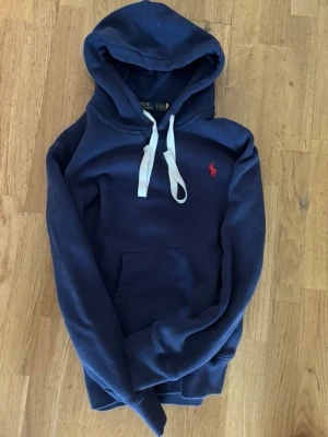 Marinblå hoodie från Polo Ralph Lauren - Snygg marinblå hoodie från Polo Ralph Lauren i slim fit-modell. Har vit dragsko i huvan, känguruficka framtill och den klassiska röda loggan broderad på bröstet. Tillverkad i mjuk bomullsblandning som känns skön mot huden.