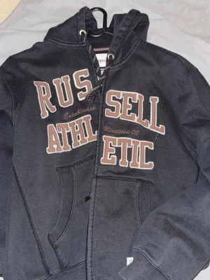 Svart hoodie från Russell Athletic - Svart hoodie med dragkedja från Russell Athletic. Stor broderad logga i beige och vitt framtill, samt känguruficka och justerbar huva med snören. Perfekt för en sportig och avslappnad stil. Den passar allt från storlek S till L beronde på hur man vill att den ska sitta.