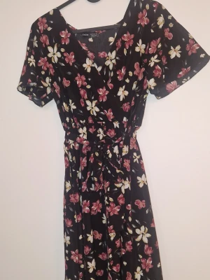 Omlottklänning  - Fin omlottklänning med v-ringning och korta ärmar, täckt av blommigt mönster. Klänningen har ett knytband i midjan som ger en snygg siluett. Perfekt för sommarens alla tillfällen.