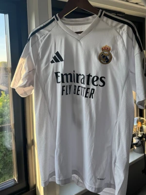 Real Madrid tröja Mbappé 9 - Säljer en vit Real Madrid matchtröja från Adidas med svarta detaljer. Tröjan har klubbmärke och Adidas-logga på bröstet, sponsortryck 'Emirates Fly Better' samt namn och nummer 'Mbappé 9' på ryggen. Tillverkad i lätt och ventilerande material. Storlek Medium. Mindre flaws vid tvätt och användning visade på bilderna.