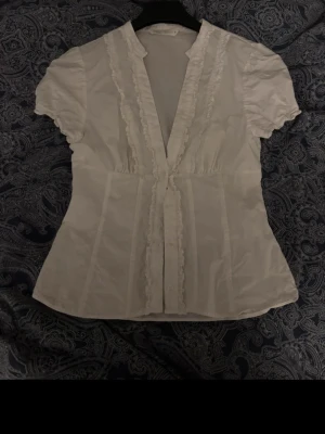 Vit blus med volangdetaljer - Söt vit blus med korta ärmar och volangdetaljer längs knäppningen framtill. Den är tyvärr för liten för mig därav sänkning. Har även små sminkfläckar på inre krage men inget som syns, går nog även att tvätta bort.