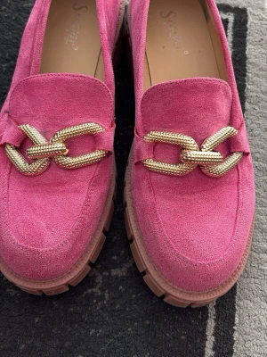 Rosa loafers med guldkedja från Seastar - Snygga rosa loafers i mockaimitation från Seastar med chunky sula och en stor guldfärgad kedjedetalj framtill. Skorna har en modern och trendig look som sticker ut och passar perfekt till dig som vill ha något extra i din outfit.