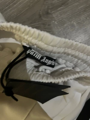 Vita shorts från Palm Angels - Snygga vita shorts från Palm Angels med elastisk midja och svarta sidopaneler. Perfekta för en avslappnad och trendig stil. Materialet känns mjukt och har en lätt glansig finish. Klassisk passform med sportig vibe.