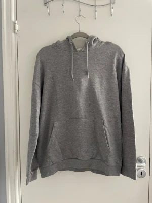 Grå basic hoodie med ficka - En enkel och stilren grå hoodie med huva och dragsnören. Hoodien har en stor magficka framtill och ribbade muddar vid ärmar och nederkant. Perfekt för en avslappnad och bekväm stil. Materialet ser ut att vara mjukt och skönt, troligen bomullsblandning.
