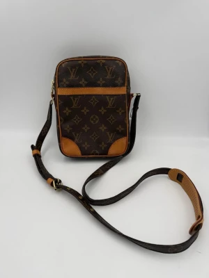 Louis Vuitton axelväska - Snygg brun axelväska från Louis Vuitton med klassiskt monogrammönster och detaljer i ljust brunt läder. Väskan har justerbar axelrem och en rektangulär form, perfekt för att bära det viktigaste. Ikonisk design som sticker ut.
