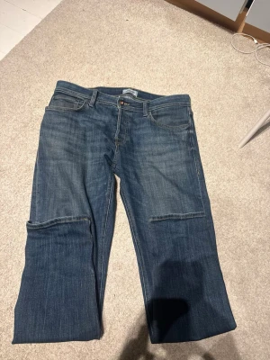 Blå raka jeans med patchdetaljer - Säljer ett par blå jeans med raka ben och tydliga patchdetaljer över knäna. Klassisk femficksmodell med normal midja och gylf med knapp. Jeansen har en avslappnad passform och är tillverkade i denim. Perfekta för en casual och trendig look.