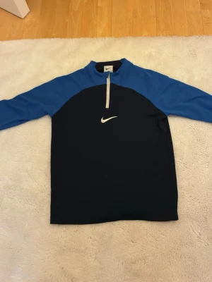 Blå och svart Nike träningströja - Snygg långärmad träningströja från Nike i blått och svart med halv dragkedja framtill och vit Nike-logga på bröstet. Perfekt för löpning eller gym. Tillverkad i ett stretchigt funktionsmaterial som andas. Pris kan diskuteras vid snabb affär!