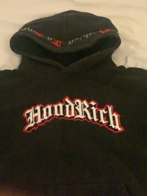Svart hoodie från Hoodrich XL - Svart hoodie från Hoodrich med stor logga i vit och röd text på bröstet. Klassisk huva med tryck på kanten och avslappnad passform. Perfekt för dig som gillar streetwear och vill sticka ut med en bold look. XL för barn. 