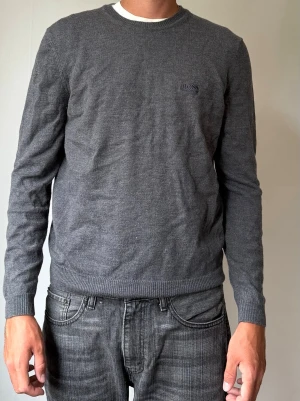 Mörkgrå stickad tröja från Hugo BOSS - Mörkgrå stickad tröja från BOSS med diskret logga på bröstet. Tröjan har rund halsringning, långa ärmar och ribbade muddar vid ärmslut och nederkant. Perfekt för en stilren och enkel look. Krympt i tvätten 
