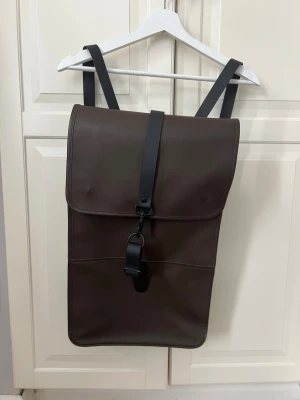 RAINS Backpack W3  - Snygg mörkbrun ryggsäck med stilren design och svart spänne framtill. Ryggsäcken har justerbara svarta axelband och en slät baksida med dragkedjeficka. Tillverkad i ett vattenavvisande material som ger en modern look.