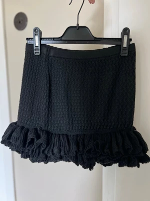 Svart stickad kortkjol med volang - Supersnygg svart stickad kortkjol med volangkant nedtill. Kjolen har en strukturerad yta och elastisk midja som ger en bekväm passform. Perfekt att styla med en enkel topp eller oversized tröja.
