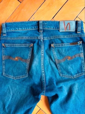 Blå jeans från Nudie Jeans - Säljer ett par klassiska blå jeans från Nudie Jeans i 100% ekologisk bomull. Jeansen har raka ben, tydliga orange sömmar och ikoniska bakfickor med vågigt broderi. Perfekta för dig som gillar stilrena och hållbara jeans. Mått 📏bredd 78cm total längd 107cm bredd vid benen 15cm.