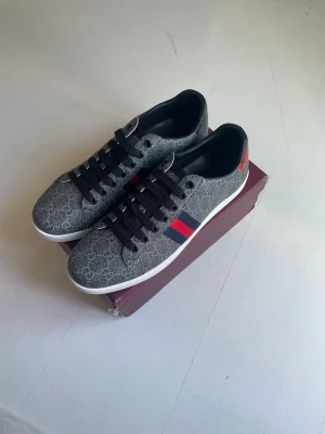 Sneakers med monogram och metallic - Snygga GG ace sneakers i lågt modell med grått monogrammönster, svarta skosnören och klassiska röd-blå ränder på sidan. Baksidan har metallicdetaljer i rött och blått med Gucci-logga. Yttermaterial i textil och läder för en lyxig känsla.
