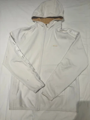Vit hoodie från BOSS med dragkedja - Snygg vit hoodie från BOSS med dragkedja framtill och huva med beige insida. Ärmarna har vita band med BOSS-logga längs sidorna. Perfekt för en clean och stilren look. Mjukt material och två fickor framtill.