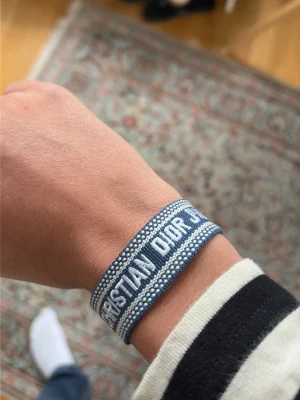 Blått Christian Dior tygarmband - Säljer ett blått tygarmband från Christian Dior med vit brodyr och texten 'CHRISTIAN DIOR' på framsidan. Armbandet har justerbara band med tofsar i ändarna och en cool, avslappnad look. Perfekt accessoar för att lyfta din outfit.