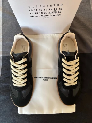 Maison Margiela svarta sneakers - Svarta sneakers från Maison Martin Margiela med beige snörning och klassisk gummisula. Ovandelen är i en mix av läder och mocka, med diskreta detaljer och vit insida. Snygg och stilren design som passar till många outfits.