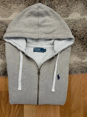 Grå zip hoodie från Polo Ralph Lauren - Snygg grå hoodie med dragkedja från Polo Ralph Lauren. Klassisk design med vit dragsko i huvan, två fickor fram och den ikoniska blå loggan broderad på bröstet. Tillverkad i mjuk bomullsblandning för skön komfort.