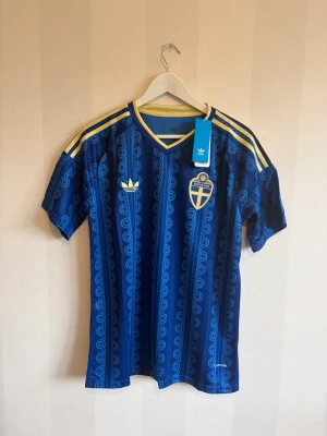 Blå Sverige fotbollströja Adidas - Säljer en blå fotbollströja från Adidas med Sveriges landslagsemblem på bröstet. Tröjan har gula detaljer, klassiska Adidas-ränder på axlarna och ett mönster i blått. Perfekt för dig som vill stötta Sverige på match eller träning.