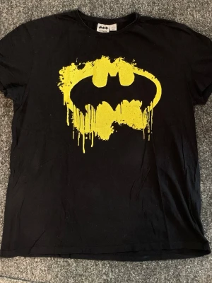 Svart Batman t-shirt med gul logga - Cool svart t-shirt från Batman med en stor gul Batman-logga framtill i en splash-effekt. Klassisk rund hals och korta ärmar. Perfekt för dig som gillar superhjältar och vill sticka ut med en ikonisk design.