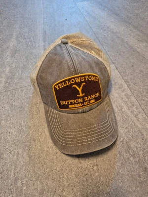 Grå truckerkeps Yellowstone Ranch - Snygg grå truckerkeps med mesh baktill och broderad patch framtill med texten 'Yellowstone Dutton Ranch Montana'. Böjd skärm och justerbar passform. Kepsen är från märket Beechfield i USA.