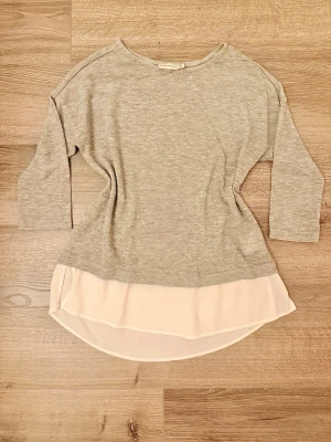 Grå topp med chiffongkant Angelology XS - Säljer en ljusgrå topp från Angelology i storlek XS. Toppen har trekvartsärm och en snygg, lös passform. Nederdelen är dekorerad med en ljusbeige chiffongkant som ger en extra touch. Defekter förekommer, se bilder.