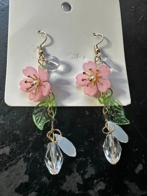 Blommiga örhängen med pärldetaljer - Unika hängande örhängen med rosa blommor, gröna blad och klara pärlliknande detaljer. Örhängena har guldfärgade krokar och inslag av transparenta och vita pärlor som ger en lekfull och färgglad look. Perfekta för dig som gillar statement-accessoarer med naturinspirerad design.