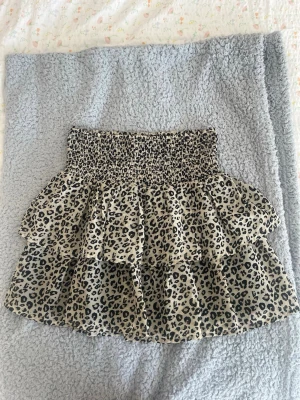 Leopardkjol med volanger - Säljer en kort kjol med leopardmönster i svart och beige. Kjolen har bred smock i midjan och dubbla volanger. Köpt i storlek 146/152 men passar mig som har storlek xs. Kan finnas några lösa trådar💗