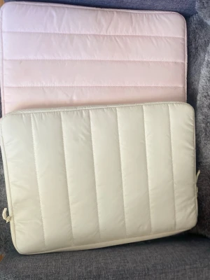 Två quiltade laptopfodral - Säljer två quiltade laptopfodral, ett i ljusrosa och ett i vitt. Båda är rektangulära med dragkedja och sydda i vadderat tyg. Perfekta för att skydda din dator i väskan eller på språng.
