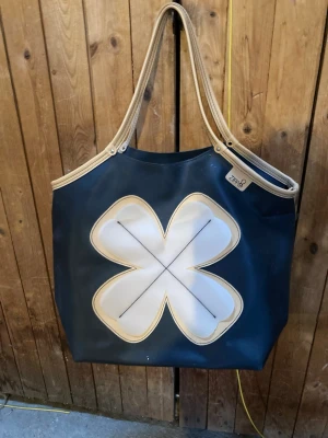 Stor shoppingväska med fyrklöver - Snygg shoppingväska i mörkblått med stort vitt fyrklövermotiv på framsidan. Väskan har beige detaljer och dubbla handtag i beige. Tillverkad i ett tåligt material som liknar plast eller återvunnet segeltyg. Perfekt för att bära mycket och sticker ut med sin coola design.