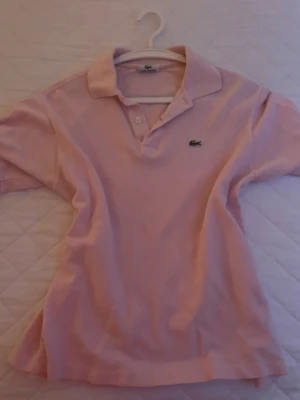 Ljusrosa pikétröja från Lacoste - Säljer en ljusrosa pikétröja från Lacoste med klassisk krage och knappar framtill. Tröjan har korta ärmar och den ikoniska krokodilloggan på bröstet. Perfekt för en sportig och stilren look.