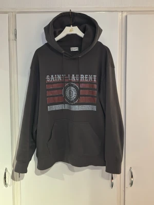 Svart Saint Laurent hoodie med tryck - Cool svart hoodie från Saint Laurent med stort tryck framtill i vitt och rött. Klassisk känguruficka och justerbar huva med snören. Mjuk bomullskänsla och avslappnad passform, perfekt för en streetwear-look.