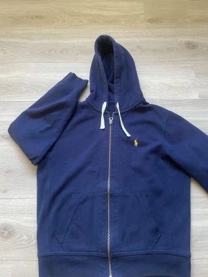 Mörkblå Ralph lauren zip hoodie med huva - Snygg mörkblå hoodie från Ralph lauren med dragkedja och huva. Har vita snören och en gul broderad logga på bröstet. Praktisk känguruficka framtill och mjukt material som känns skönt mot huden. Perfekt för en avslappnad stil. Helt ny 10/10 skick 