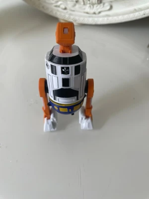 Star Wars robotfigur i plast - Cool Star Wars-inspirerad robotfigur i plast med vita, svarta, orangea och blå detaljer. Figuren har tre ben och ett cylinderformat huvud med orange topp. Perfekt för samlare eller som dekoration.