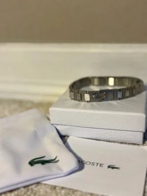 Silverfärgat armband från Lacoste - Stilrent armband i silverfärgad metall från Lacoste. Armbandet har rektangulära länkar med små mönstrade detaljer och den klassiska Lacoste-loggan på spännet. Kommer med original ask och vit tygpåse med grönt krokodilmärke.