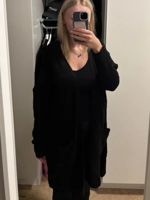 Svart lång kofta från Lager 157  - Säljer en svart lång kofta från Lager 157 i storlek XS/S. Koftan har öppen front, två stora fickor framtill och är stickad i ett mjukt material. Perfekt att slänga över en outfit för en chill look.