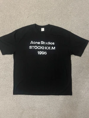 Svart Acne Studios t-shirt Stockholm - Säljer en svart t-shirt från Acne Studios med vit text 'Acne Studios STOCKHOLM 1996' på bröstet. Klassisk rak passform och rund halsringning. Perfekt för dig som gillar stilrena och ikoniska plagg.