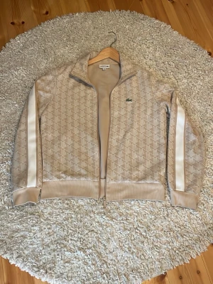 Beige track jacket från Lacoste - Snygg beige track jacket från Lacoste med diskret mönster och vita ränder längs ärmarna. Dragkedja framtill, ribbade muddar och klassisk ståkrage. Perfekt för en sportig och trendig look.