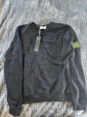 Svart sweatshirt från Stone Island - Säljer en svart sweatshirt från Stone Island med klassisk rund hals och ribbade muddar. Tröjan har den ikoniska Stone Island-loggan på vänster ärm och är tillverkad i mjukt bomullsmaterial. Perfekt för en avslappnad och stilren look.