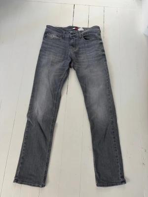 Grå Tommy Jeans Scanton Slim - Säljer ett par gråa slim fit jeans från Tommy Jeans, modell Scanton Slim. Jeansen har klassisk femficksdesign, normal midja och raka ben. Tillverkade i stretchigt denim för extra komfort. Perfekta för en avslappnad och trendig look.