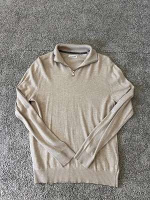 Beige halvzip tröja Jack & Jones - Snygg beige stickad tröja från Jack & Jones med halvzip och krage. Tröjan har långa ärmar, ribbade muddar och nederkant. Perfekt för lager på lager och enkel att matcha med olika outfits.