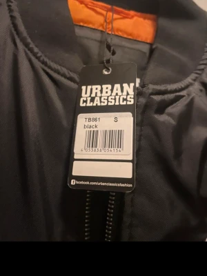 Svart bomberjacka Urban Classics Pepsi - Snygg svart bomberjacka från Urban Classics med klassisk Pepsi-logga på bröstet. Jackan har dragkedja framtill, ribbade muddar vid ärmslut och nederkant samt orange innerfoder. Perfekt för dig som gillar retrostil och streetwear.