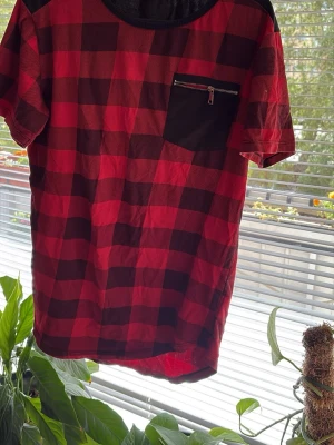 Röd och svart rutig t-shirt Charman - Säljer en röd och svart rutig t-shirt från Charman, tillverkad i Italien. T-shirten har en svart bröstficka med dragkedja och rund halsringning. Perfekt för dig som gillar streetwear och vill sticka ut lite extra.