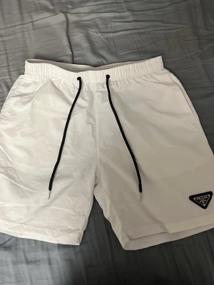 Prada badshorts - Snygga vita shorts från Prada med svart snörning i midjan och elastisk resår. Klassisk modell med fickor på sidorna och en bakficka. Perfekta för sommaren och har en stilren look med diskret Prada-logga på benet. Pris kan diskuteras vid snabb affär.