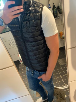 Svart lättviktsdunväst med dragkedja - Snygg svart dunväst med quiltad design och hög krage. Västen har dragkedja framtill och två sidofickor. Perfekt att slänga över en t-shirt eller hoodie för en casual look. Materialet är glansigt och lätt, vilket gör den enkel att bära och matcha.