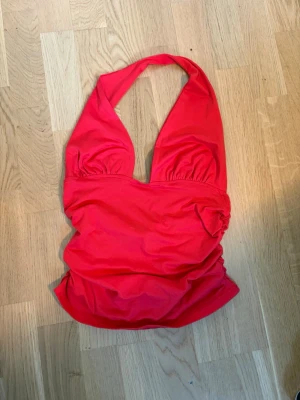 Röd halterneck baddräkt - Snygg röd baddräkt med halterneck och djup urringning. Modellen har rynkade detaljer vid bysten och är tillverkad i ett stretchigt material som passar perfekt för strand eller pool. Klassisk och trendig design som ger en cool look.