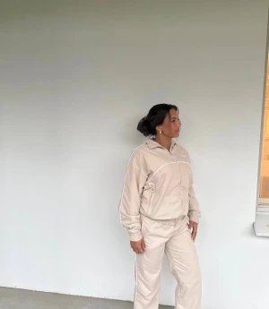 Beige tracksuit set med dragkedja - Säljer ett beige tracksuit set med matchande jacka och byxor. Jackan har dragkedja framtill, resår i ärmslut och midja samt vita detaljer längs sömmarna. Byxorna har resår i midjan och raka ben.