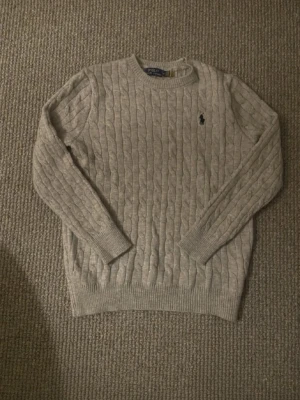 Beige kabelstickad tröja Polo Ralph Lauren - Kabelstickad beige tröja från Polo Ralph Lauren med rund halsringning och lång ärm. Tröjan har den klassiska broderade logotypen i marinblått på bröstet och ribbade muddar vid ärmslut och nederkant. Perfekt för en stilren och avslappnad look.