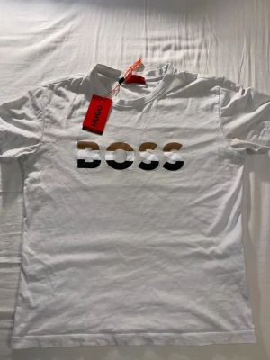 Vit Hugo Boss t-shirt med logga - Snygg vit t-shirt från Hugo Boss med stor BOSS-logga i svart, guld och vit på bröstet. Klassisk rund hals och korta ärmar. Perfekt basplagg med stilren design och skön passform. Tillverkad i mjuk bomull för maximal komfort.