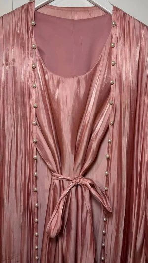 Abaya 🌸 - Säljer en lång rosa Abaya  i glansigt material med knyt i midjan och dekorativa pärlor längs kanterna. Klänningen har långa ärmar och ett elegant fall som ger en lyxig känsla.